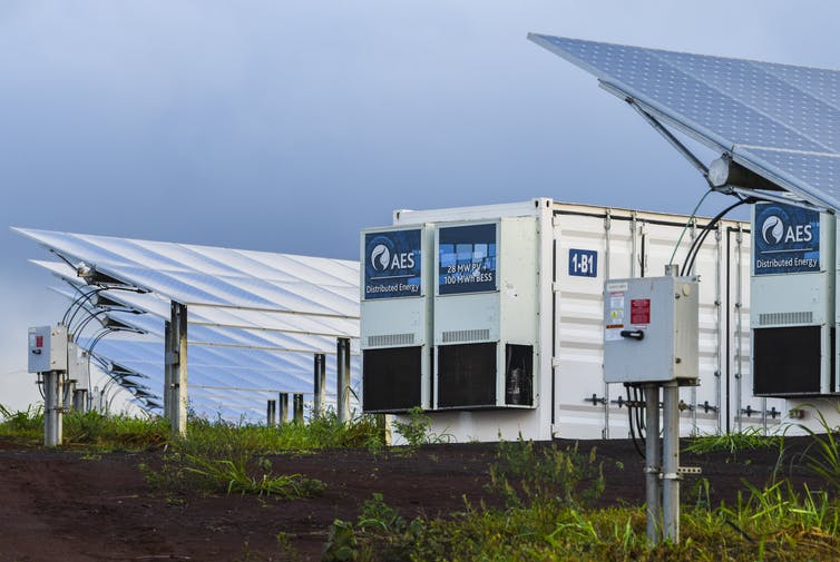 Rows of solar panels and two batteries the size of small shipping containers sit in a field.